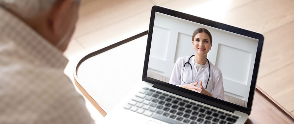 Over-the-shoulder view of a senior man having a video call with a doctor, whose image is displayed on a laptop screen.