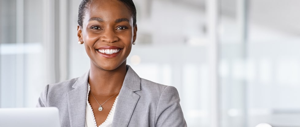 A professional woman in a gray suit sitting at a desk, smiling warmly, in a modern office environment.